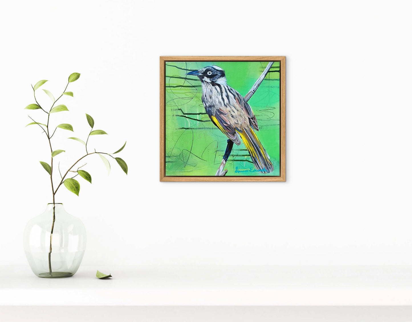 Framed artwork of a bird on a green background next to a vase with a plant on a white surface