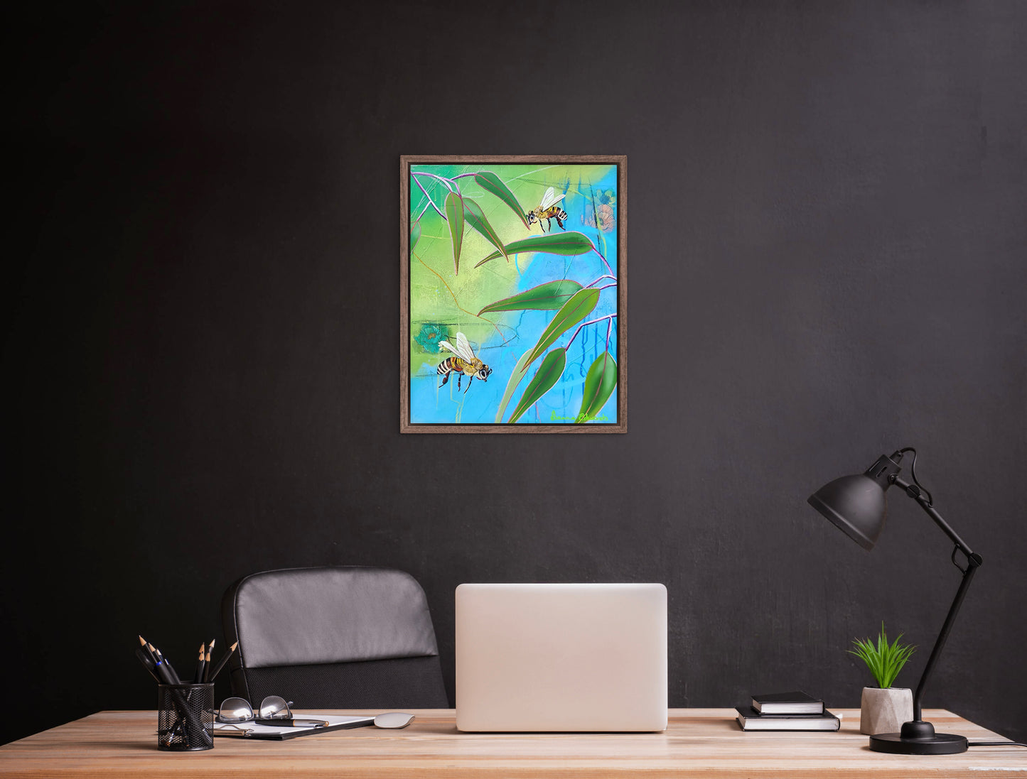 Framed artwork of bees on leaves above a desk with a laptop and lamp.