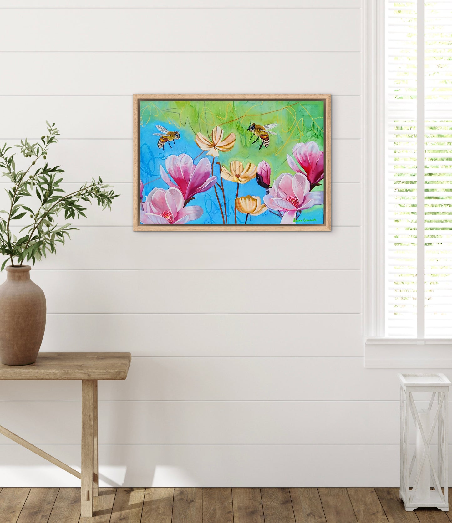 Framed artwork of flowers and bees on a white wall with a wooden table and plant.