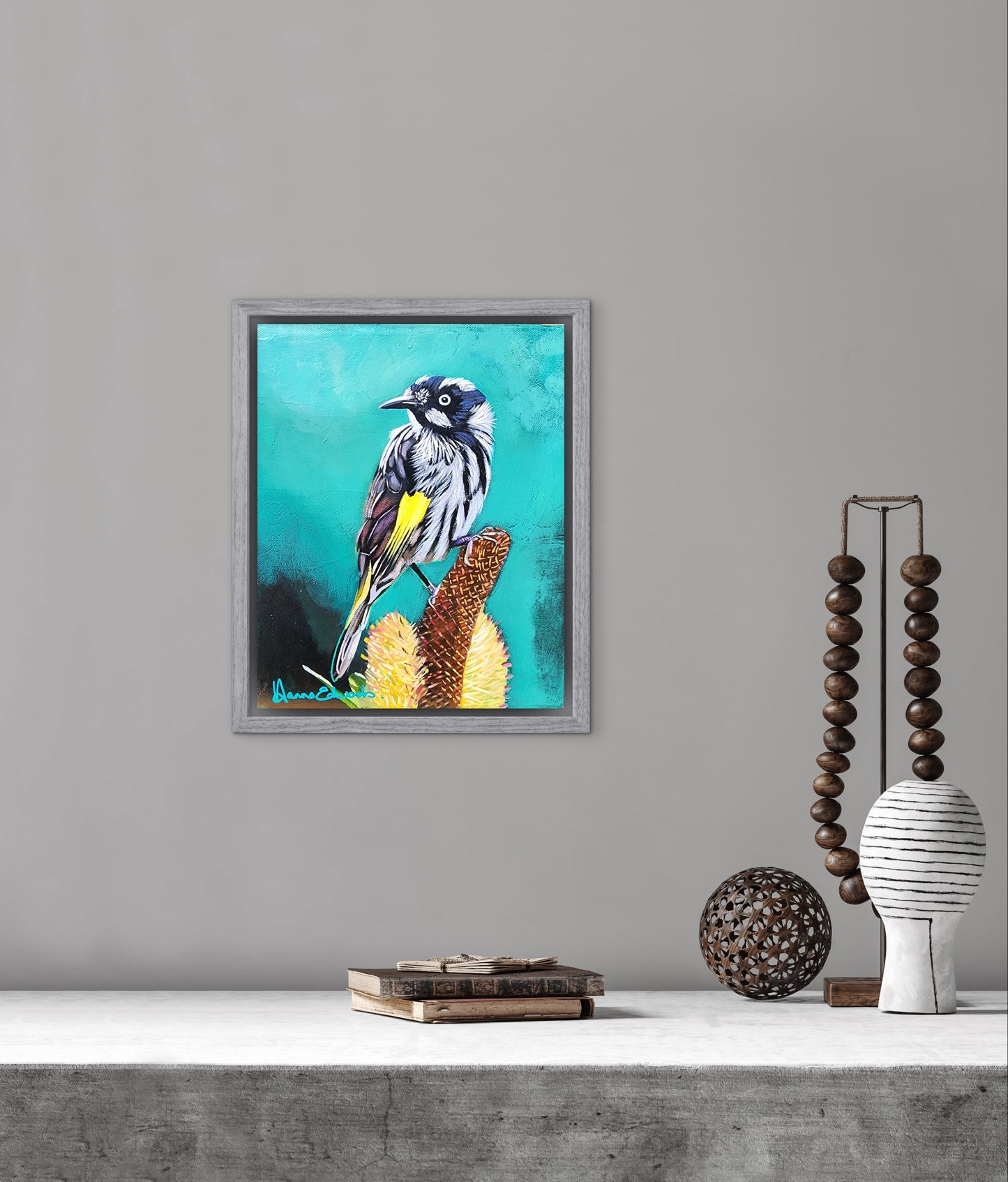 Framed artwork of a bird on a branch with a teal background, displayed on a wall above a shelf.