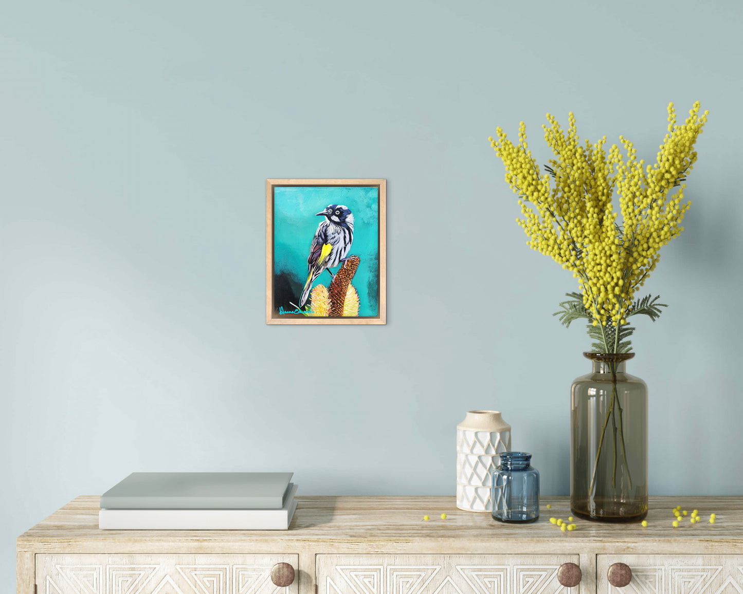 Decorative setting with a framed artwork of a bird, a vase with yellow flowers, and books on a wooden surface against a light blue wall.