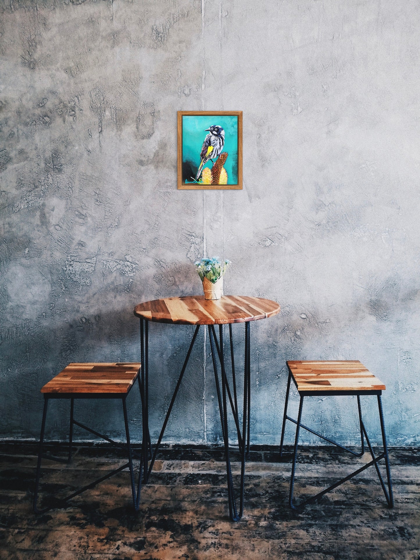 Wooden table with two stools against a textured gray wall with a framed picture.