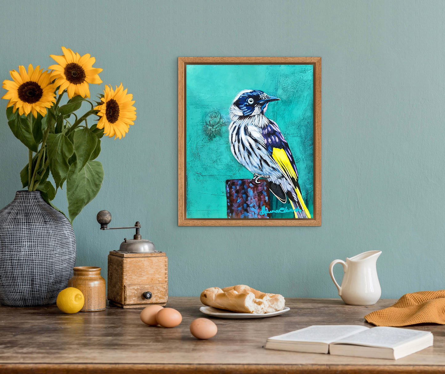 Framed bird artwork on a wall above a wooden table with sunflowers, a pitcher, and other items.