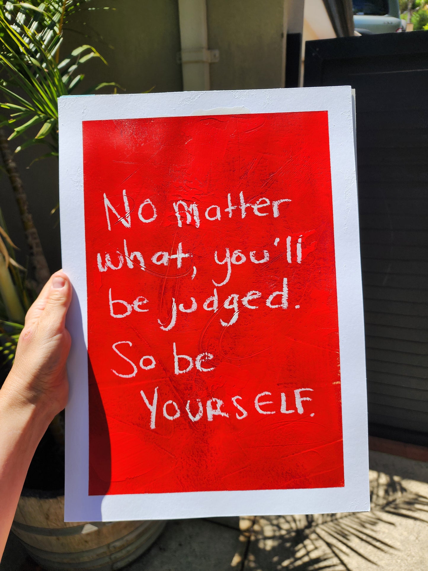 Hand holding a red painting with white quote No matter what, you'll be judged. So be yourself.