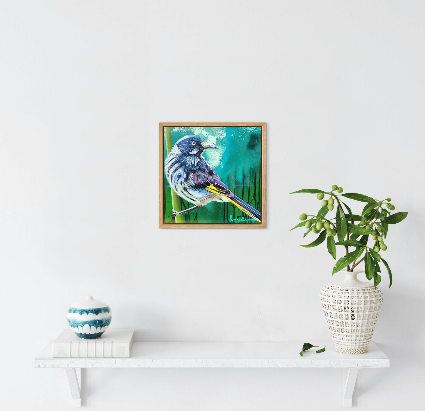 Framed artwork of a bird on a white wall with a plant and decorative items below.