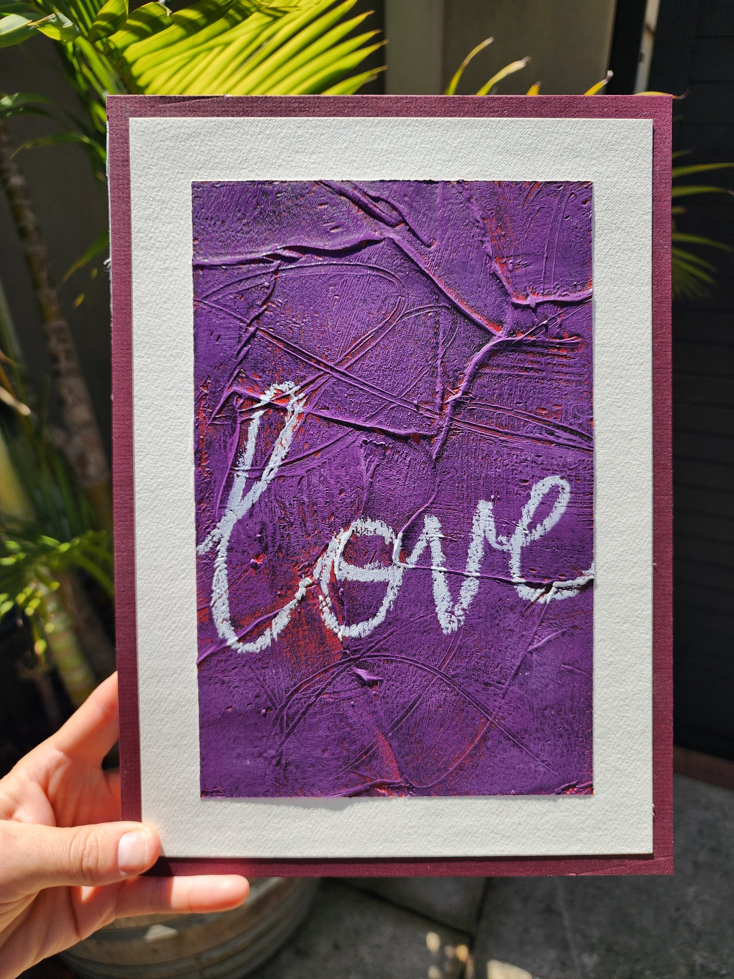 Heavily textured painting in purple with pops of red coming through with the word Love close up with a white boarder glued to a red/violet matt board held outside