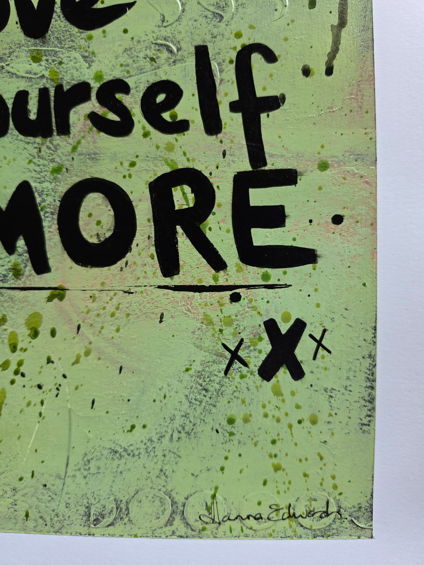 A close-up of a mixed-media art piece featuring the word 'MORE' in bold black lettering on a textured light green background with black ink splatters and scribbles.