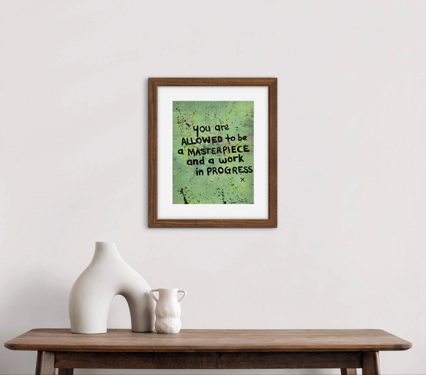 A framed artwork with a motivational quote hangs on a white wall staged to see how it looks above a wooden console table with white ceramic vases. The print features a textured green background with black text reading 'you are ALLOWED to be a MASTERPIECE and a work IN PROGRESS'.