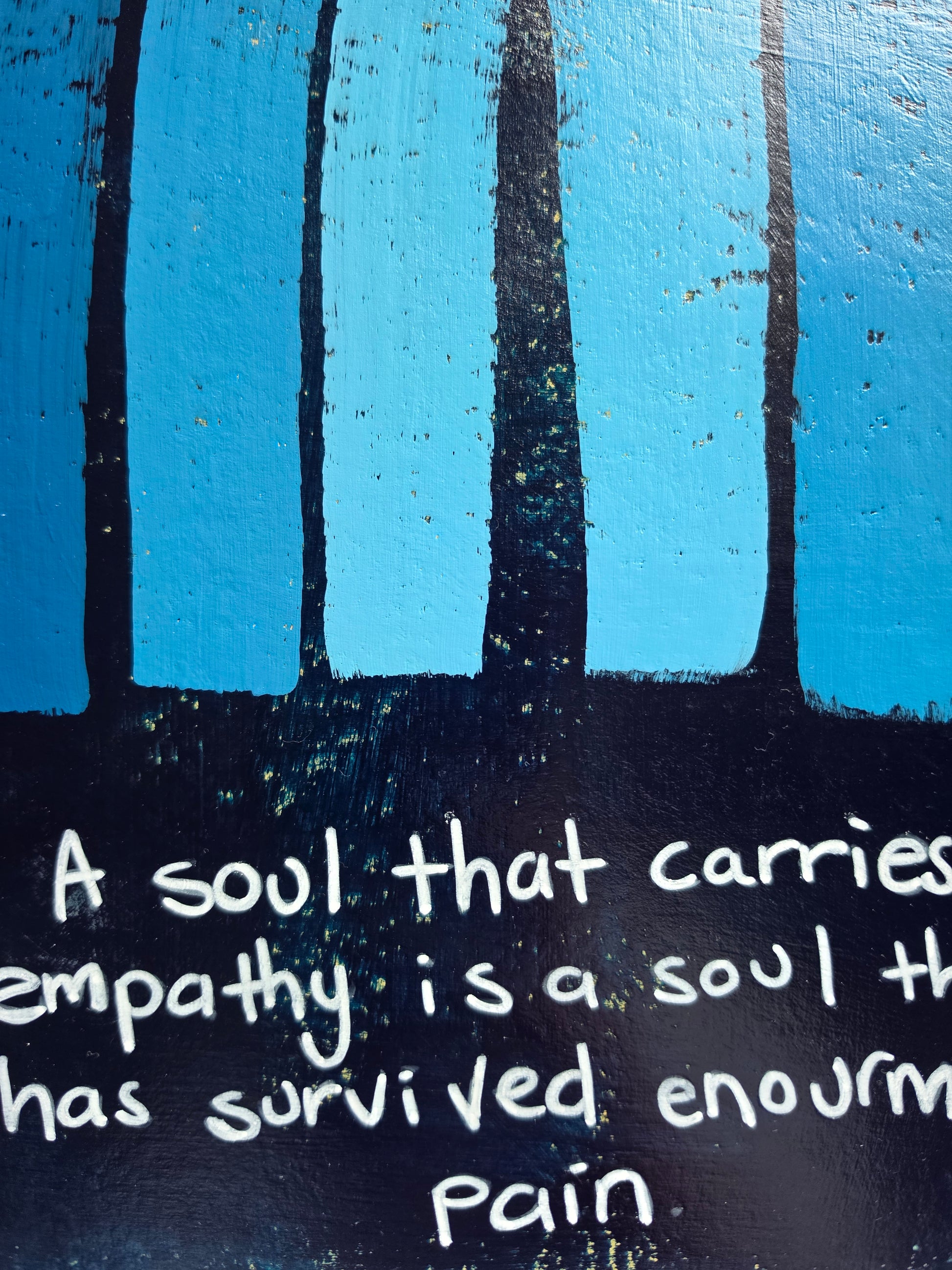 A close-up of a painted A blue painting with pops of yellow undernesth with a white border featuring a blue and black textured rainbow design and a handwritten quote, "A soul that carries empathy is a soul that has survived enormous pain"