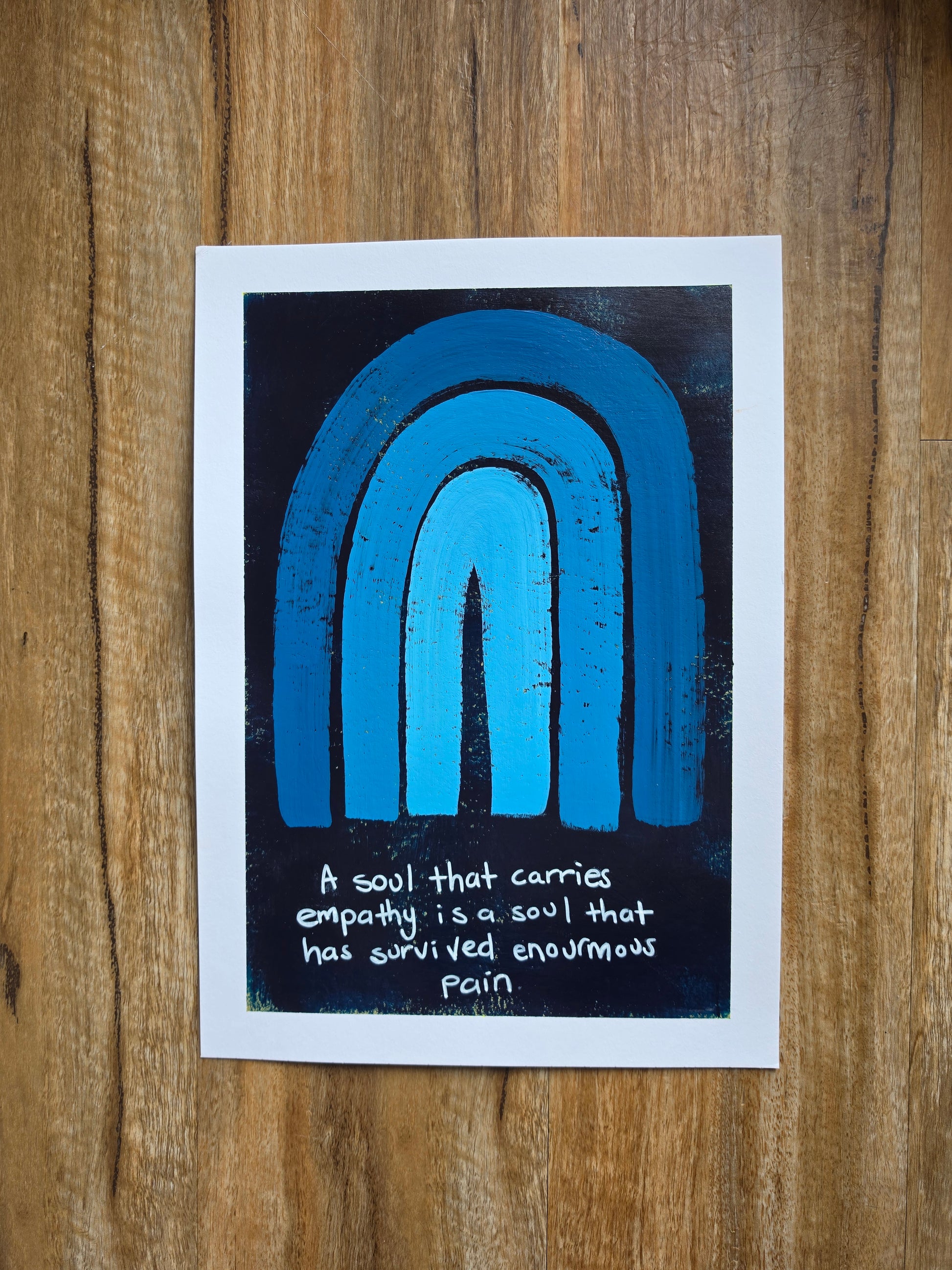 A blue painting with pops of yellow undernesth with a white border featuring a blue and black textured rainbow design and a handwritten quote, "A soul that carries empathy is a soul that has survived enormous pain"