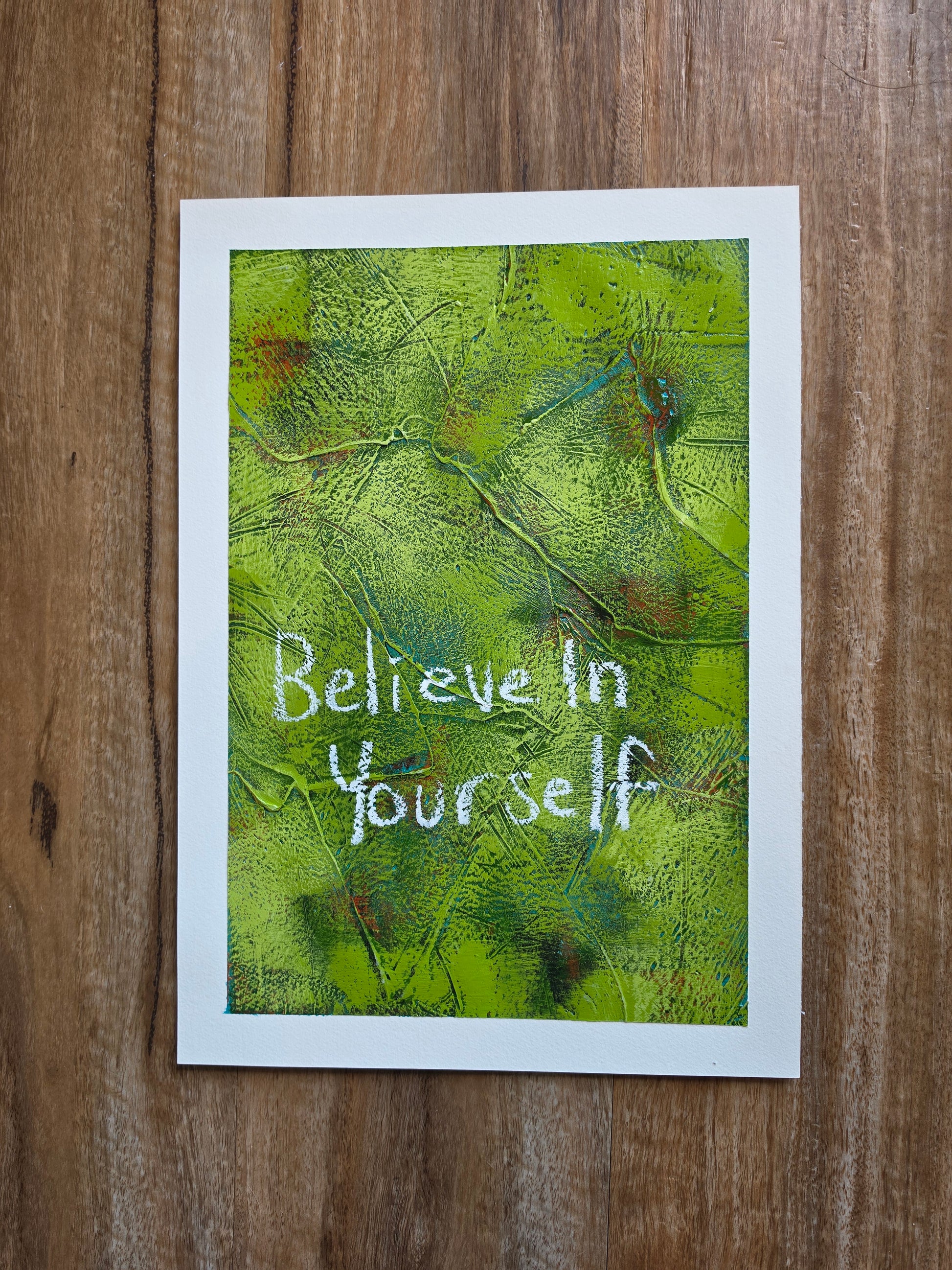 Abstract painting in green with green and red pops of colour in underneath layers with words Believe in yourself in white handwriting with alot of texture