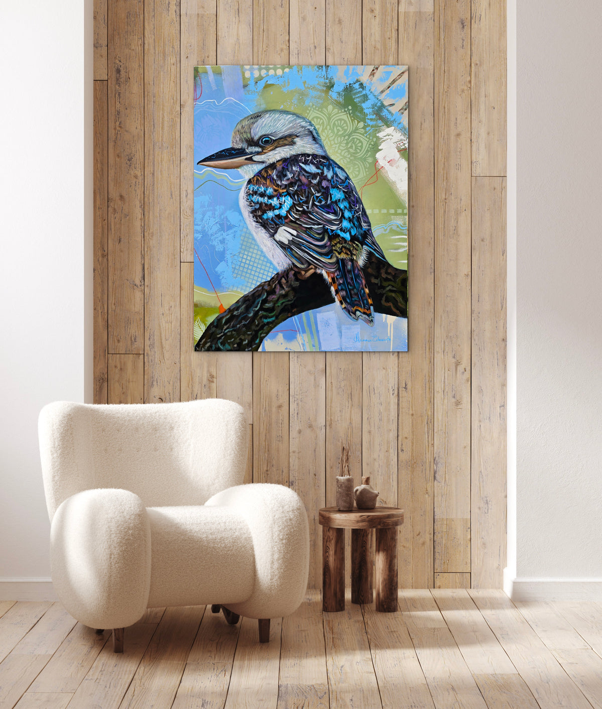 “Liberation” original kookaburra painting by Australian artist Hanna Edwards, displayed on a timber-panelled wall above a cream armchair in a contemporary interior, showing how the colourful artwork looks in a living space.