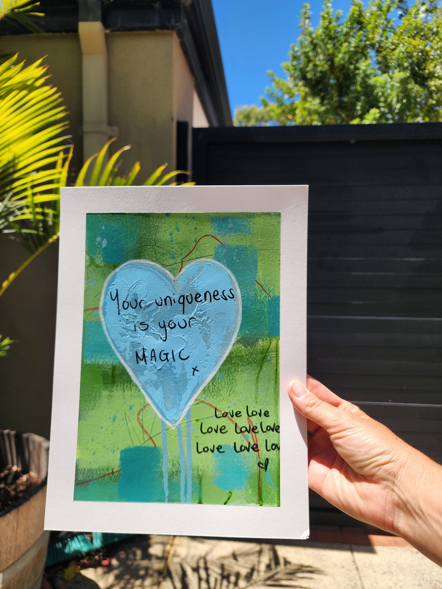 artwork with a blue heart and text held by a hand outdoors.