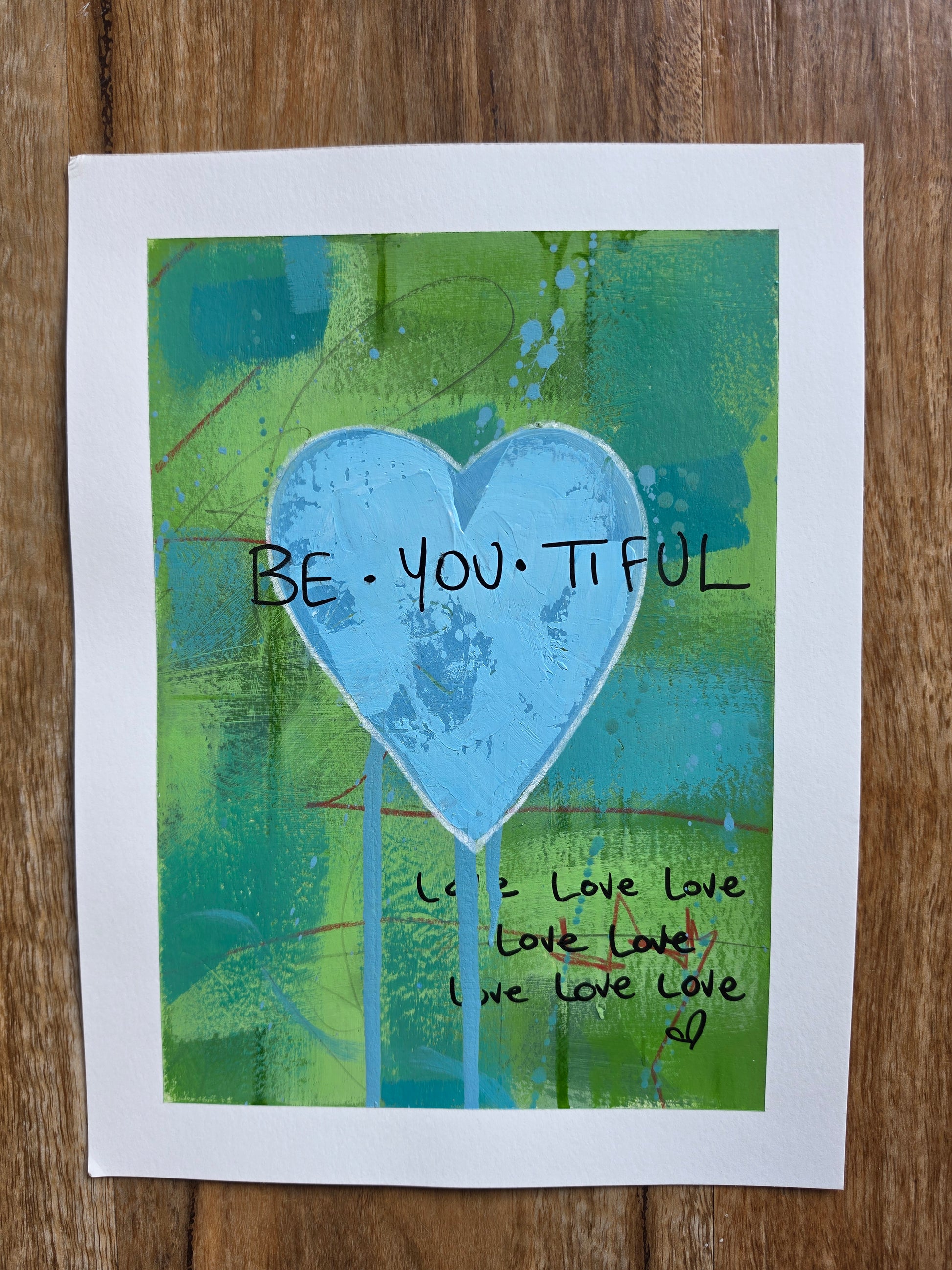 Abstract artwork with a blue heart and 'BE YOU TIFUL' text on a wooden surface