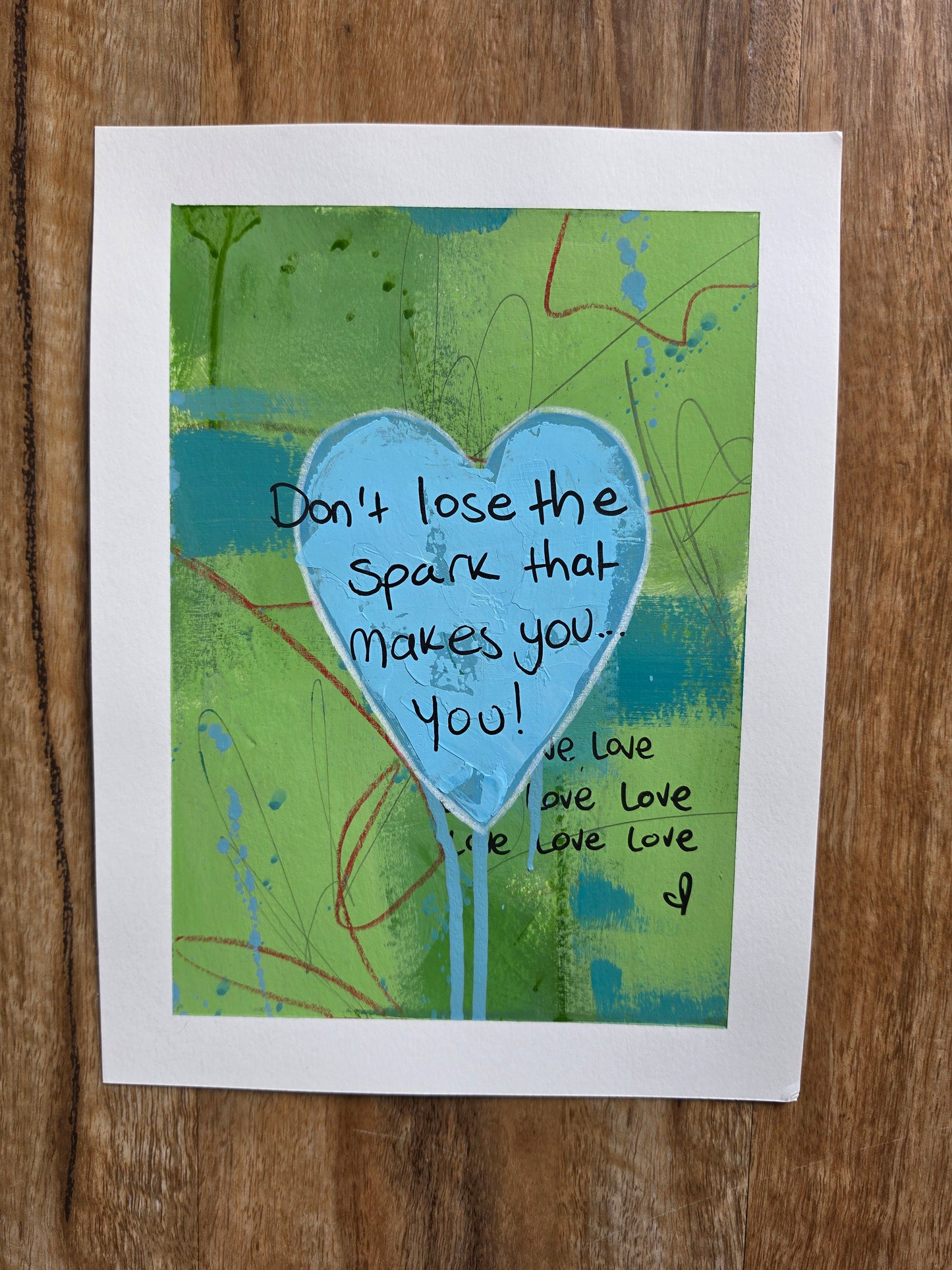 Abstract artwork with a blue textured heart and text on a green background, placed on a wooden surface.