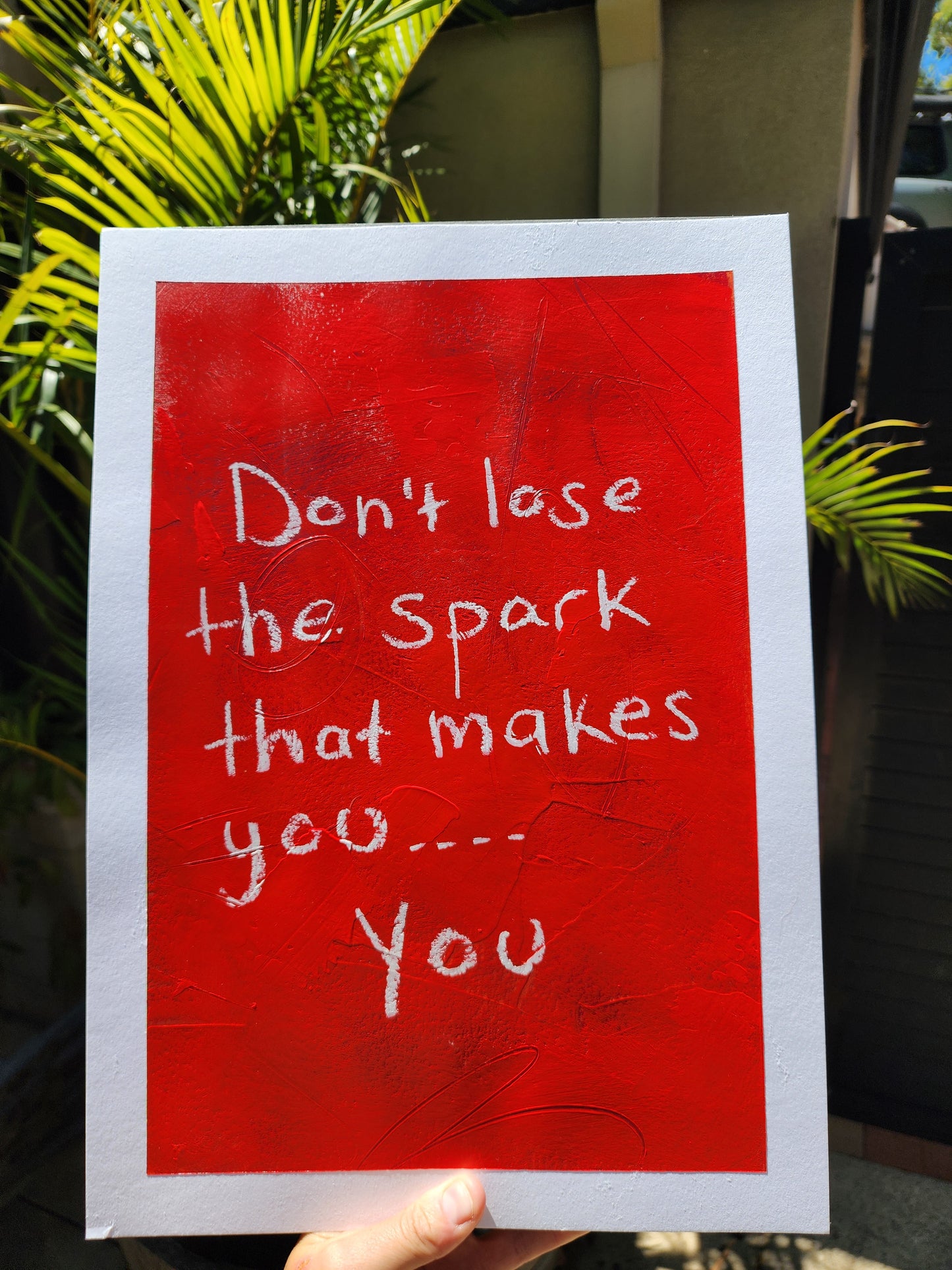 Red painting with white text 'Don't lose the spark that makes you... You' held by a person outdoors.