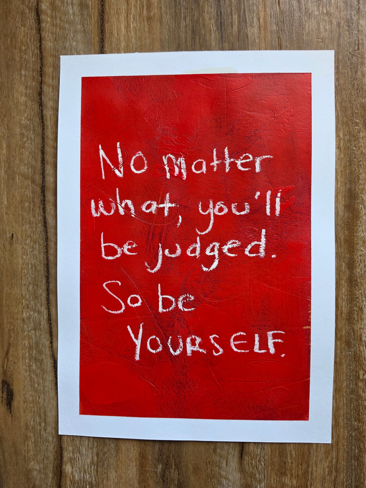 Red painting with white inspirational quote No matter what, you'll be judged. So be yourself. On a wooden surface
