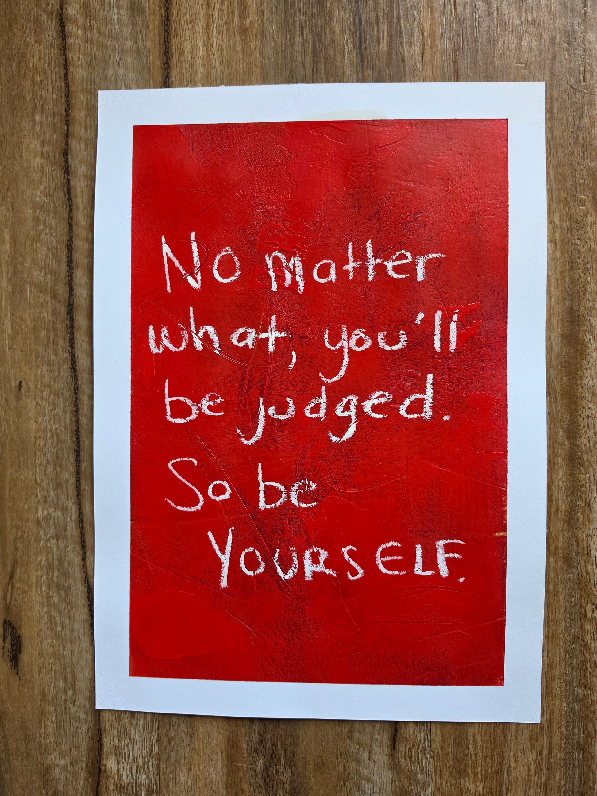 Red painting with white inspirational quote No matter what, you'll be judged. So be yourself. On a wooden surface