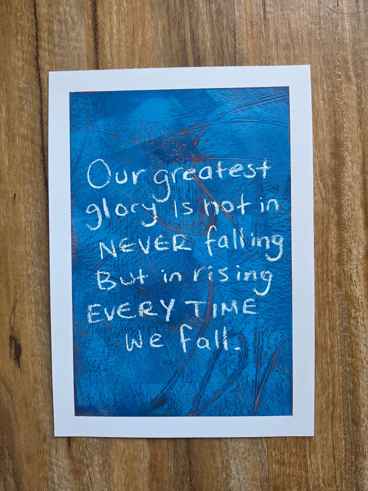 Blue painting with pops of red with white text "Our greatest glory lies not in never falling but rising everytime we fall " on a wooden surface