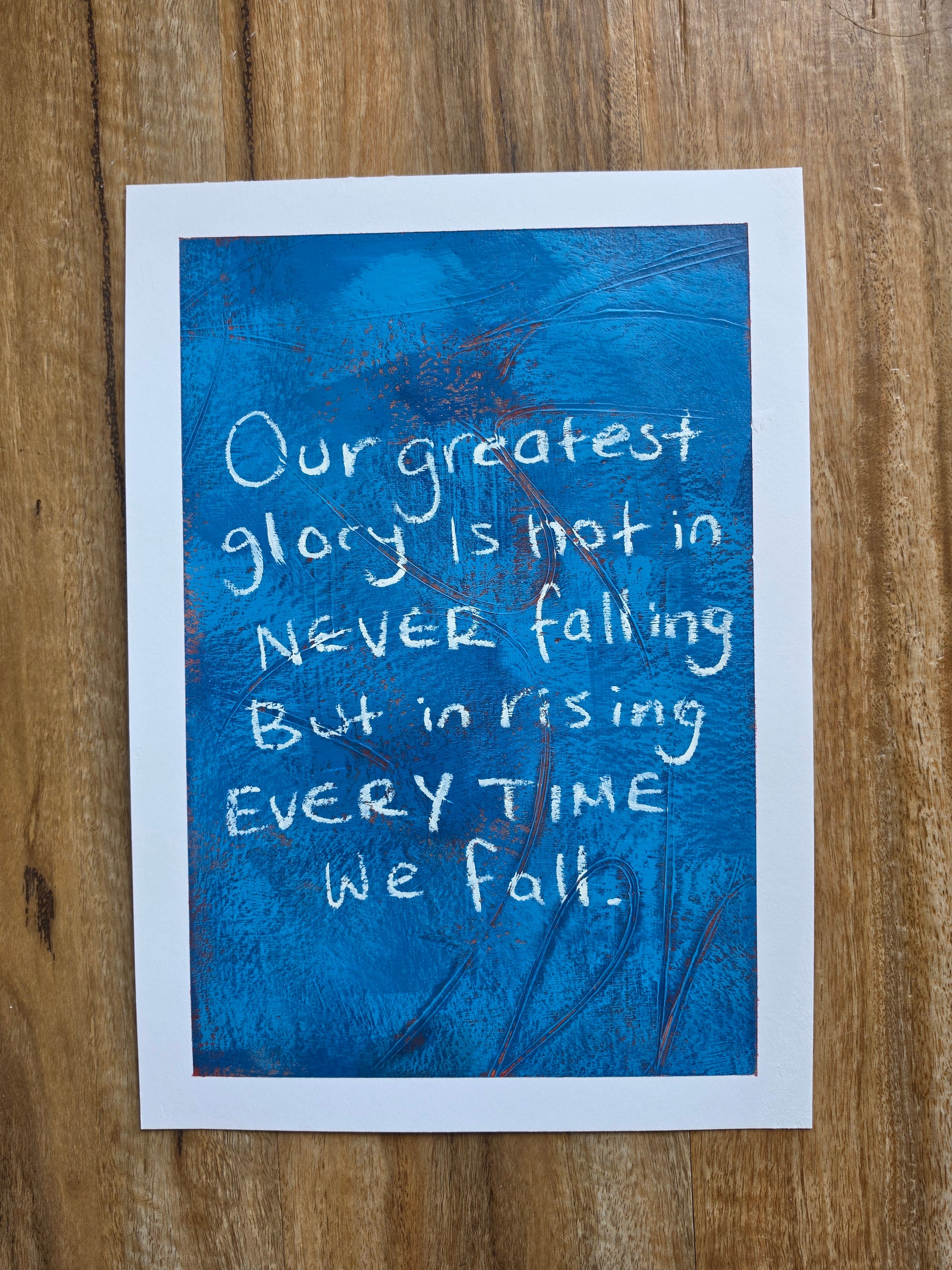 Blue painting with pops of red with white text "Our greatest glory lies not in never falling but rising everytime we fall " on a wooden surface