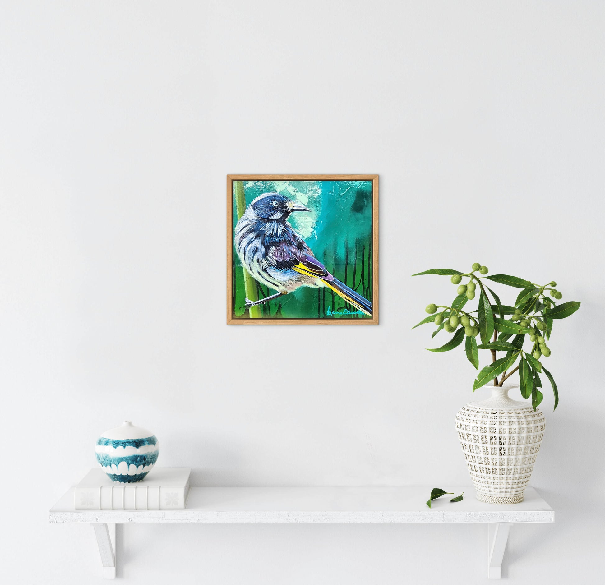 Framed artwork of a bird on a white wall with a plant and decorative items below.