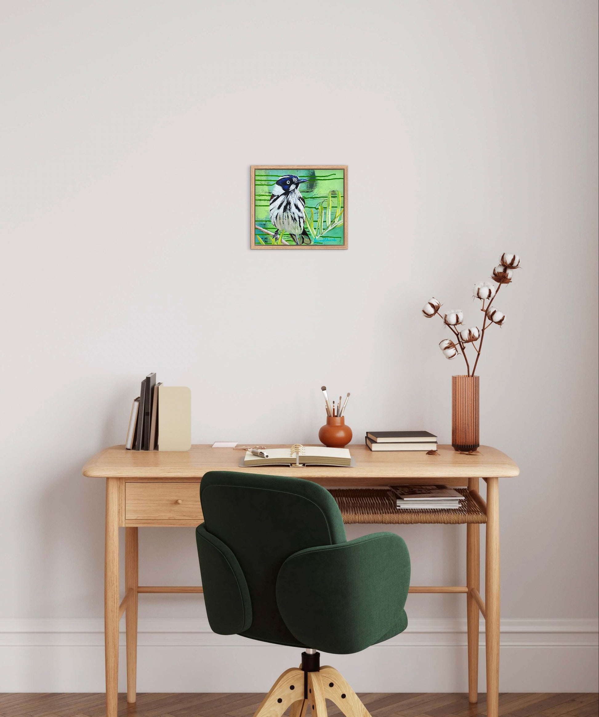 Wooden desk with green chair, books, and a vase against a white wall with a framed picture.
