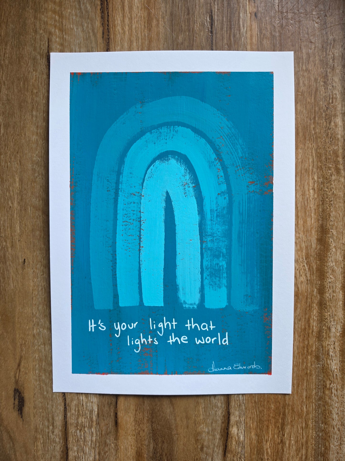 Painting with a white border a blue background with red popping through and turquoise rainbow design with the quote 'It's your light that lights the world' written by hand in white with artist Hanna Edwards signature 