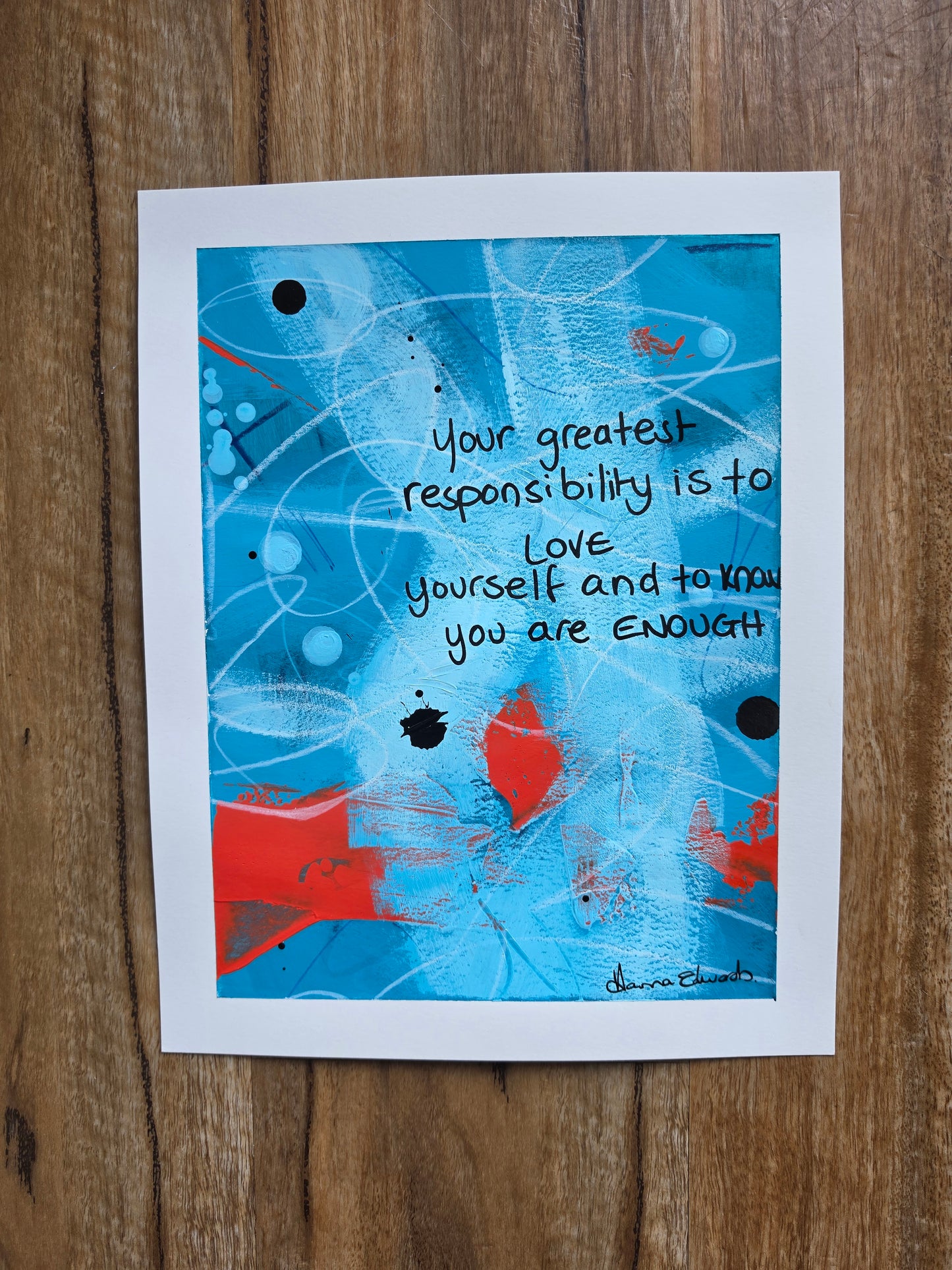  abstract art  with quote handwritten reading "Your greatest responsibility is to love yourself and know you are enough" displayed flat on a wooden surface. The artwork features a white border with a blue, red, and black abstract design and handwritten text.