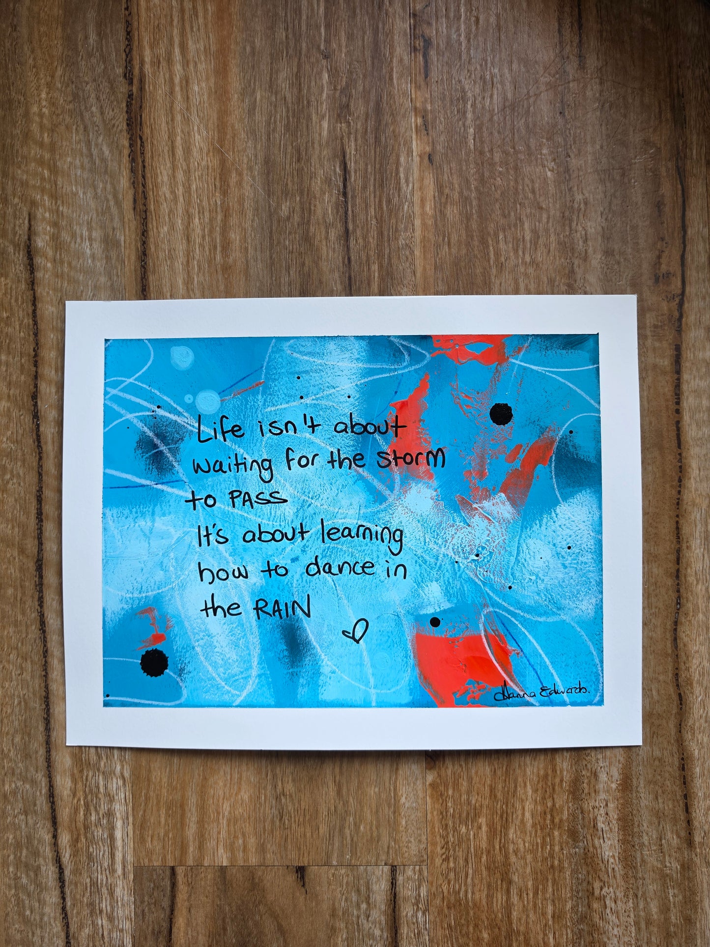 An abstract painting with the quote 'Life isn't about waiting for the storm to pass. It's about learning how to dance in the rain' by artist Hanna Edwardsh, displayed on a wooden surface.