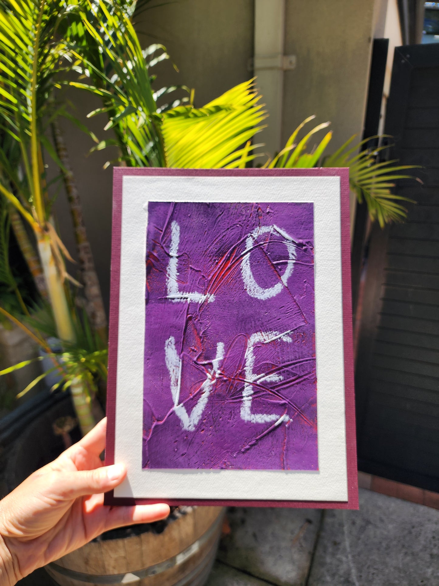 Heavily textured painting in purple with pops of red coming through with the word Love a white boarder glued to a red violet matt board