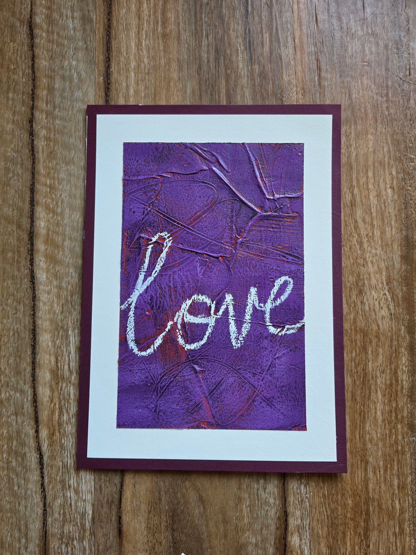 Heavily textured painting in purple with pops of red coming through with the word Love in white handwriting with a white boarder glued to a red violet matt board
