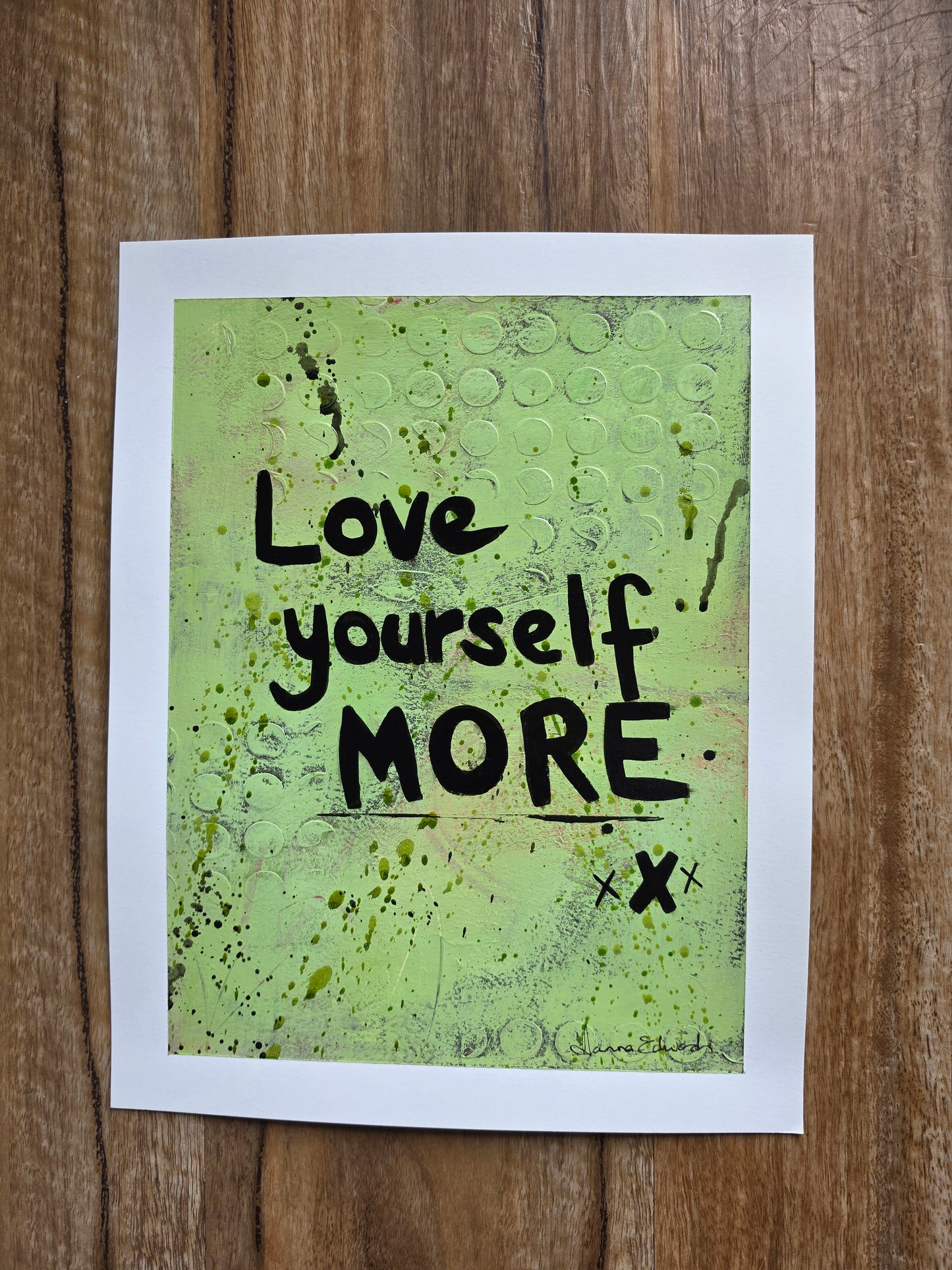 Art with a white border featuring the hand-lettered text 'Love yourself MORE' in black over a textured lime green background, displayed flat on a brown wooden surface.