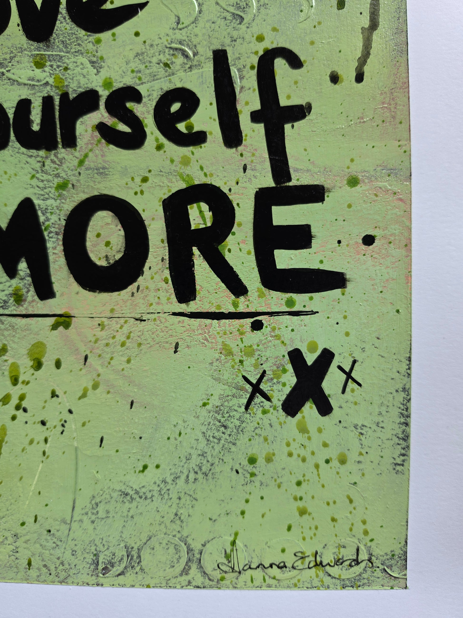 A close-up of a mixed-media art piece featuring the word 'MORE' in bold black lettering on a textured light green background with black ink splatters and scribbles.