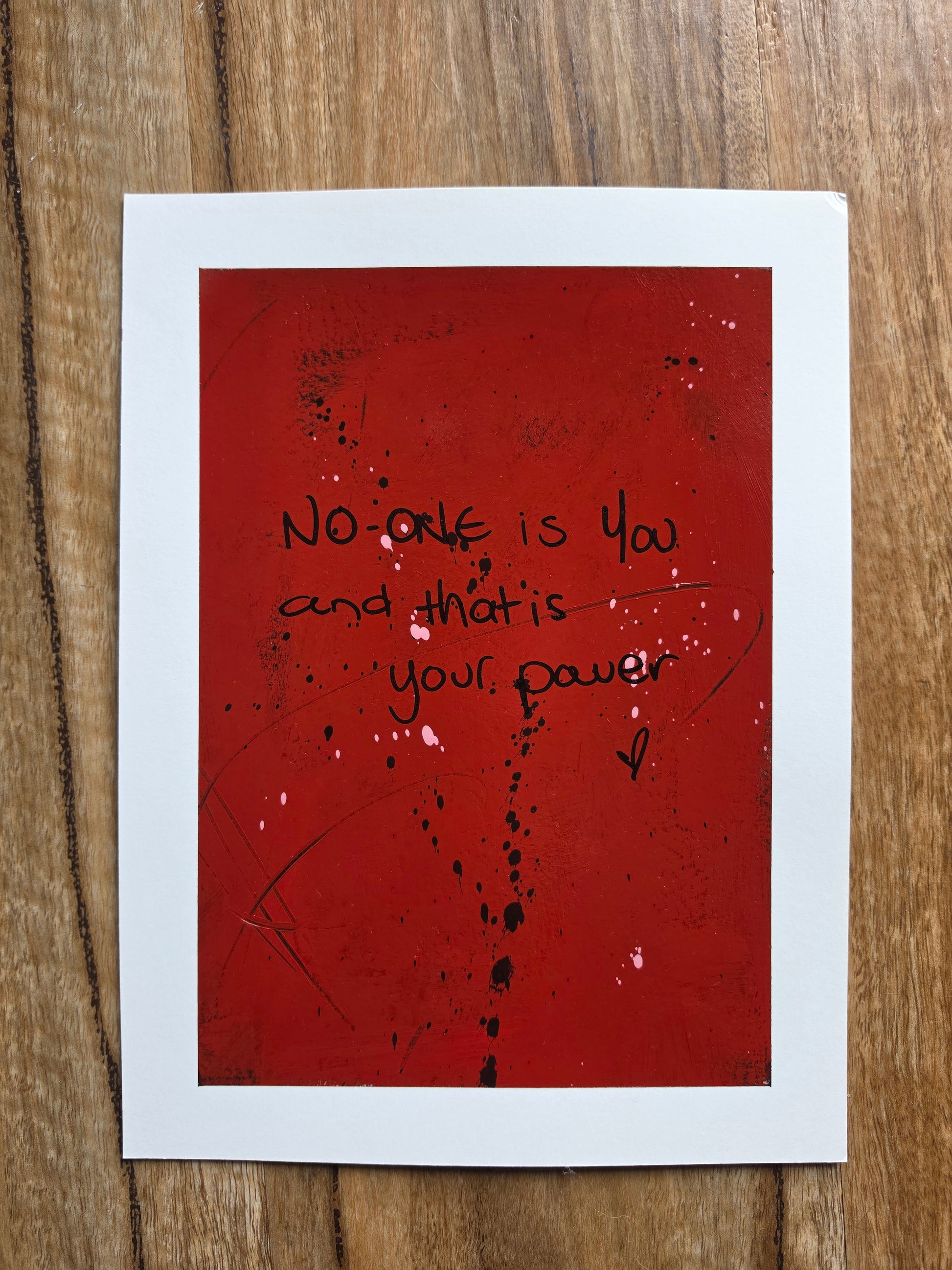 Red painting with black text No one is you and that is your power with black and pink sploges on a wooden surface