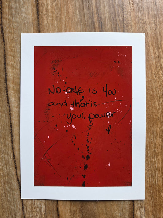 Red painting with black text No one is you and that is your power with black and pink sploges on a wooden surface