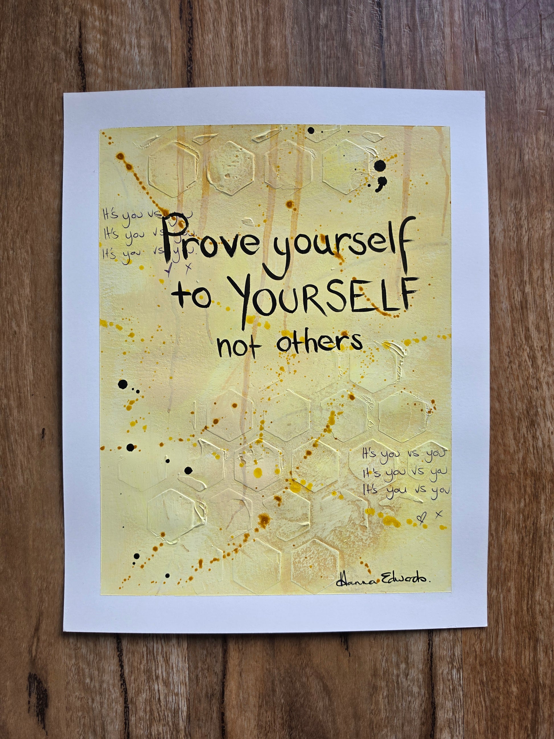 A painting featuring the quote 'Prove yourself to YOURSELF not others' by Hanna Edwards, displayed flat on a dark wood background. The piece has a white border and a central design with a textured yellow background, black lettering, and gold glitter hexagonal patterns.