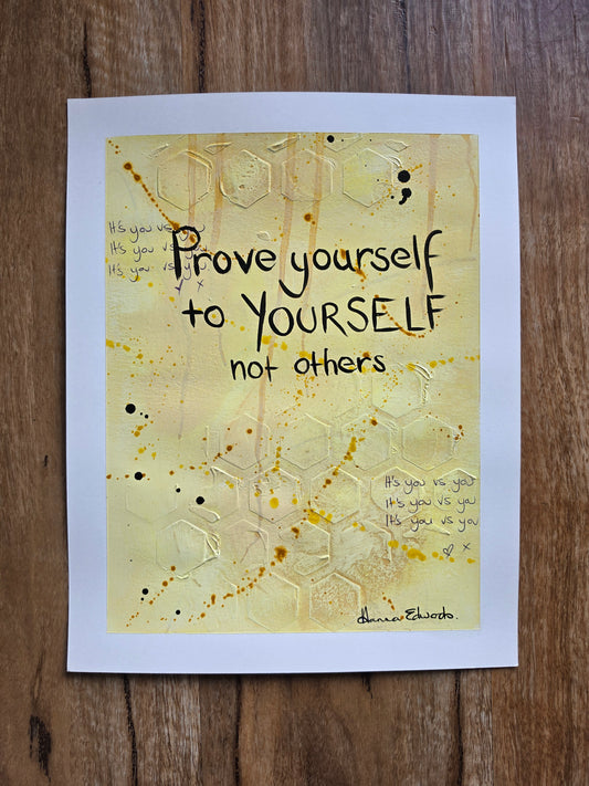 A painting featuring the quote 'Prove yourself to YOURSELF not others' by Hanna Edwards, displayed flat on a dark wood background. The piece has a white border and a central design with a textured yellow background, black lettering, and gold glitter hexagonal patterns.