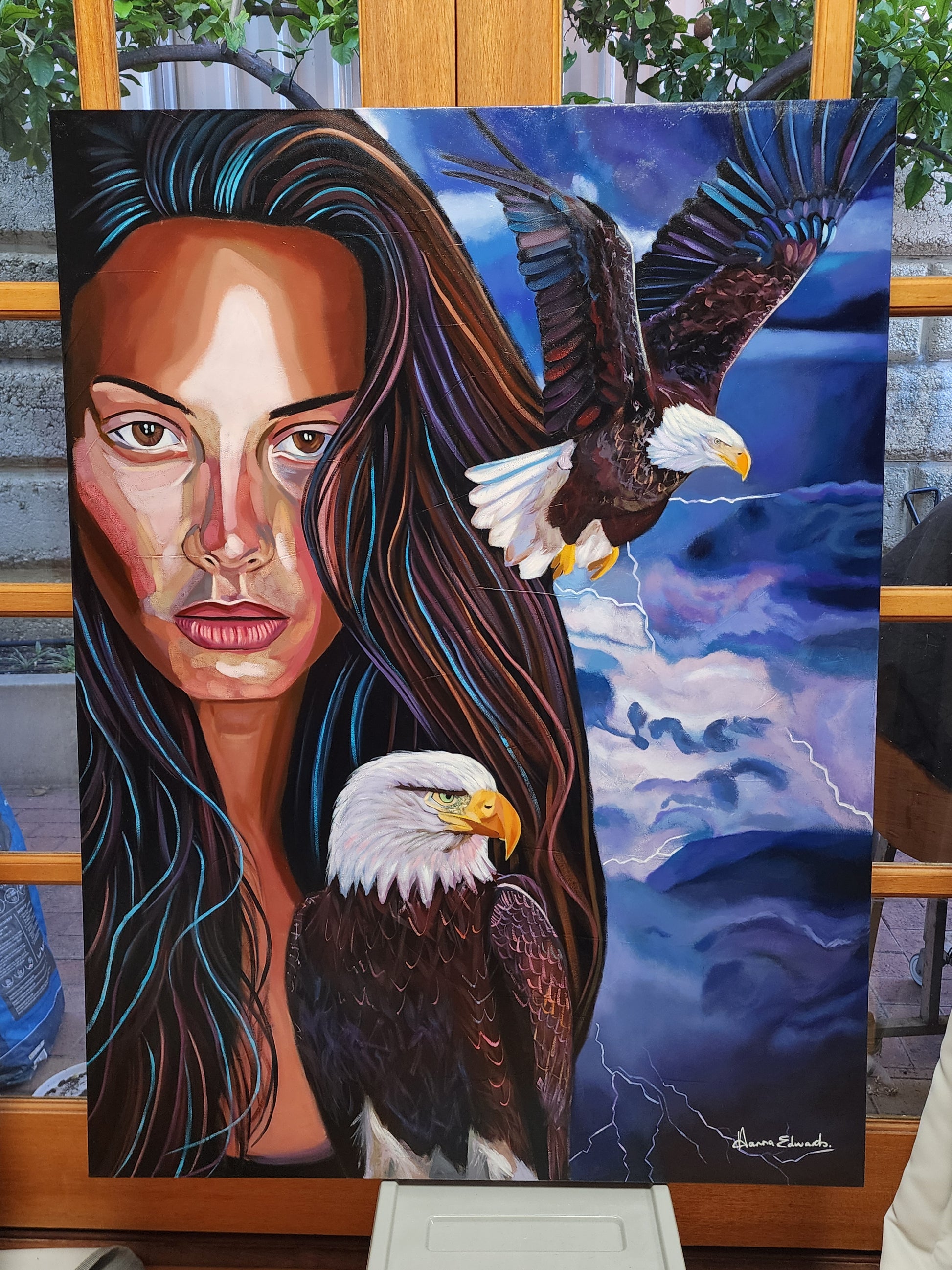 A large canvas painting displayed on an easel, featuring a woman's face with long dark hair, two bald eagles, and a stormy blue sky with lightning.