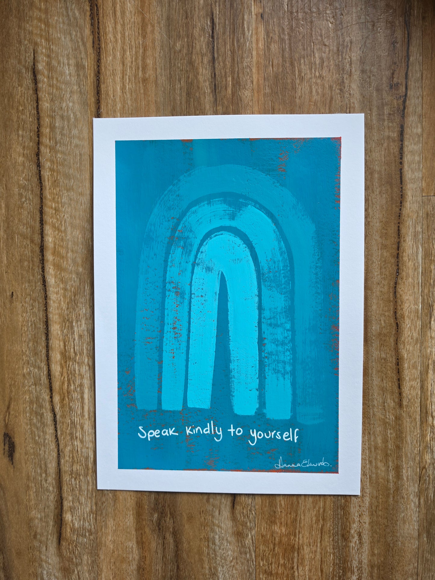 Blue painting with red popping through underneath layer with a white border featuring a textured turquoise blue abstract arch design and the handwritten quote 'Speak kindly to yourself' in white