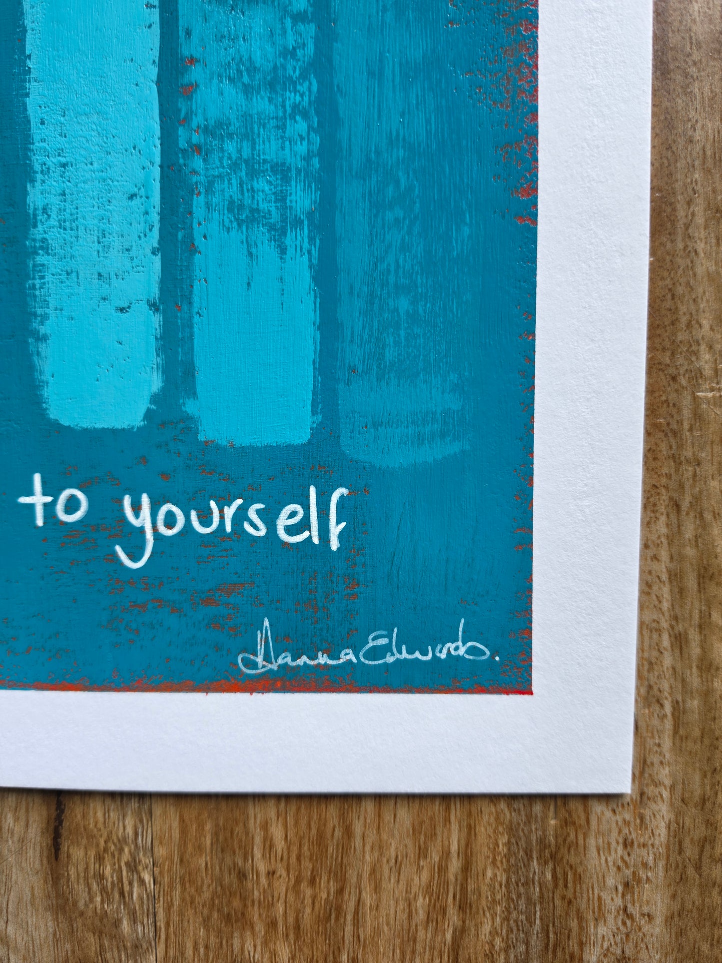 A close-up of a printed art piece on a blue and turquoise abstract design with the words ' speak kindly to yourself' with artist Hanna Edwards signature in white