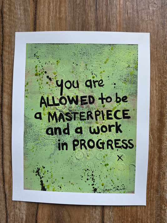 Painting with a white border lies on a wooden surface. The artfeatures a textured light green background with black hand-lettered text that reads, 'you are ALLOWED TO BE a MASTERPIECE and a work in PROGRESS'.