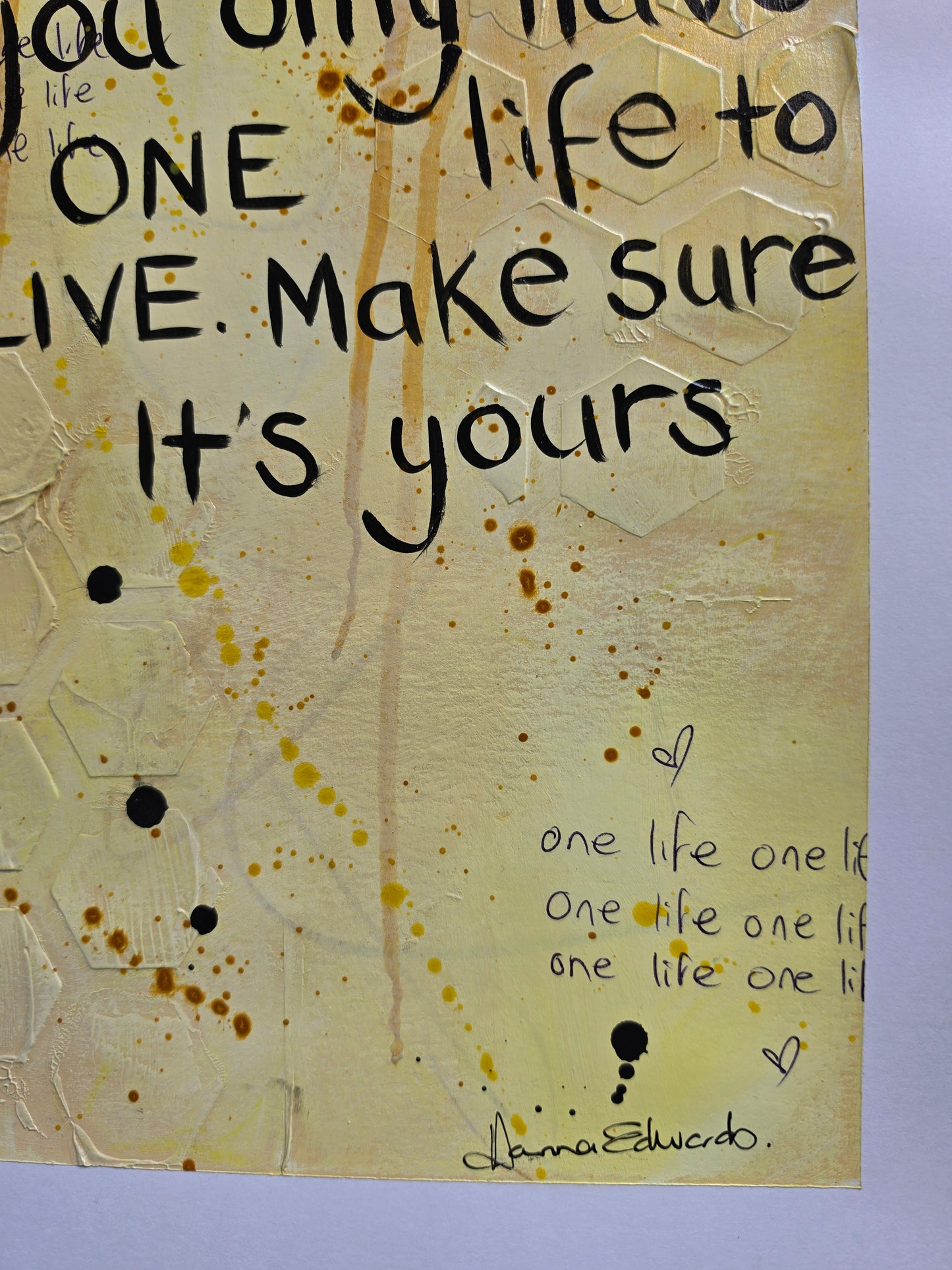 A close-up of a textured, cream-colored mixed-media art work featuring hand-lettered black text, yellow paint splatters, and a signature of artist Hanna Edwards in the bottom right corner.