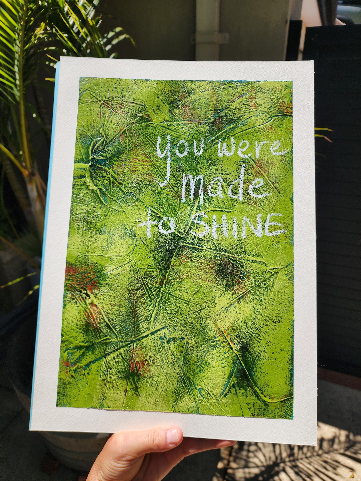 Abstract painting in green with pops of green , teal and red with the words You Were Made to Shine handwritten in white held by a hand outside