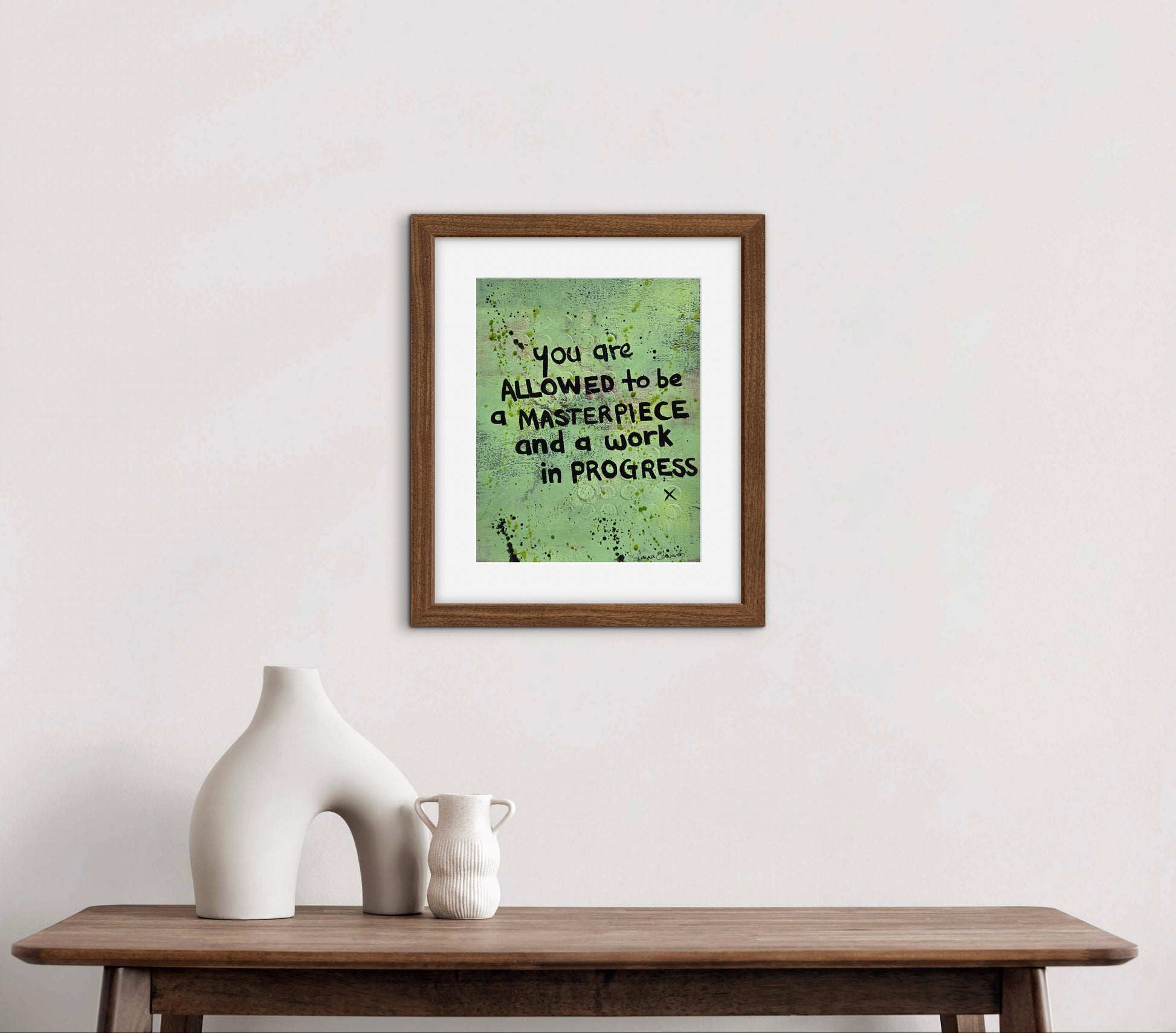 A framed artwork with a motivational quote hangs on a white wall staged to see how it looks above a wooden console table with white ceramic vases. The print features a textured green background with black text reading 'you are ALLOWED to be a MASTERPIECE and a work IN PROGRESS'.